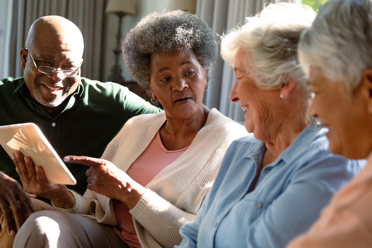 Three happy diverse senior woman and african american male friend sitting on sofa and using tablet.