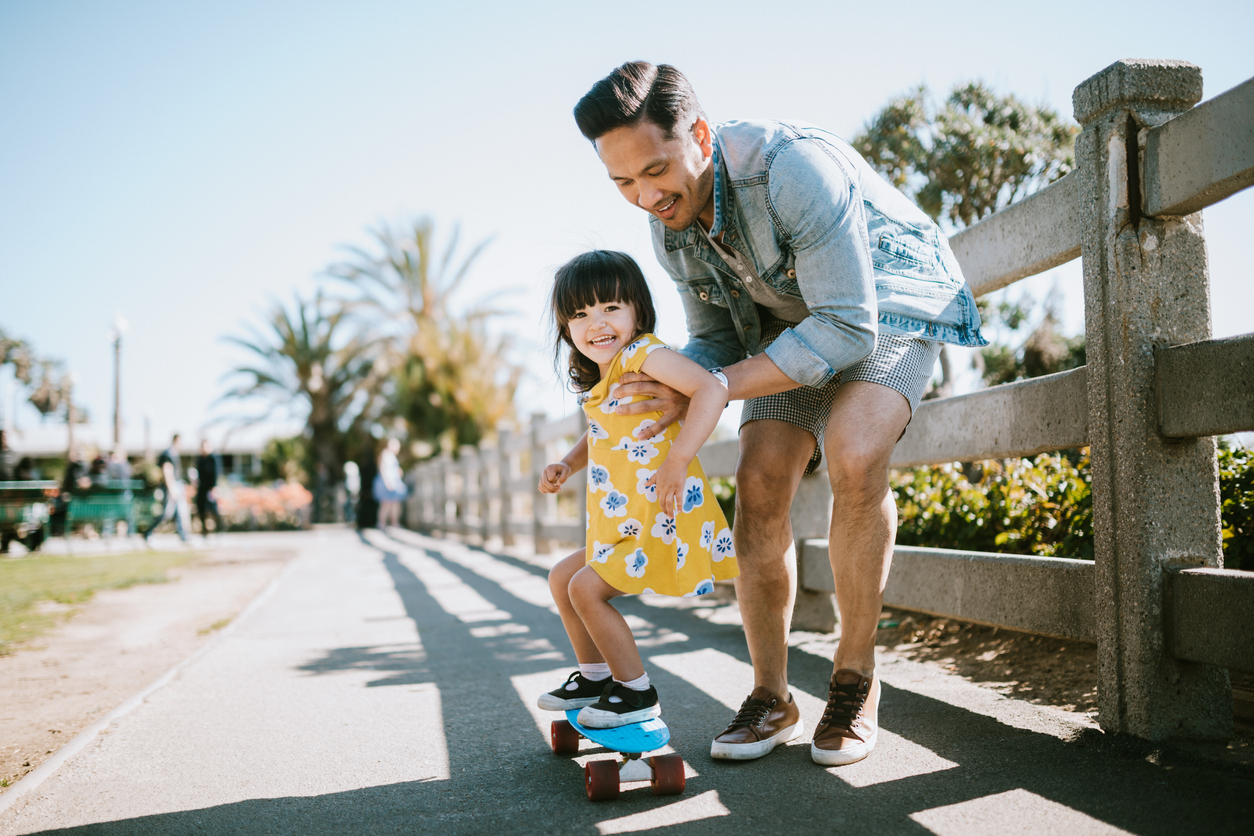 Father Helps Young Daughter Ride Skateboard A dad helps his little girl go skateboarding, holding her waist for support.